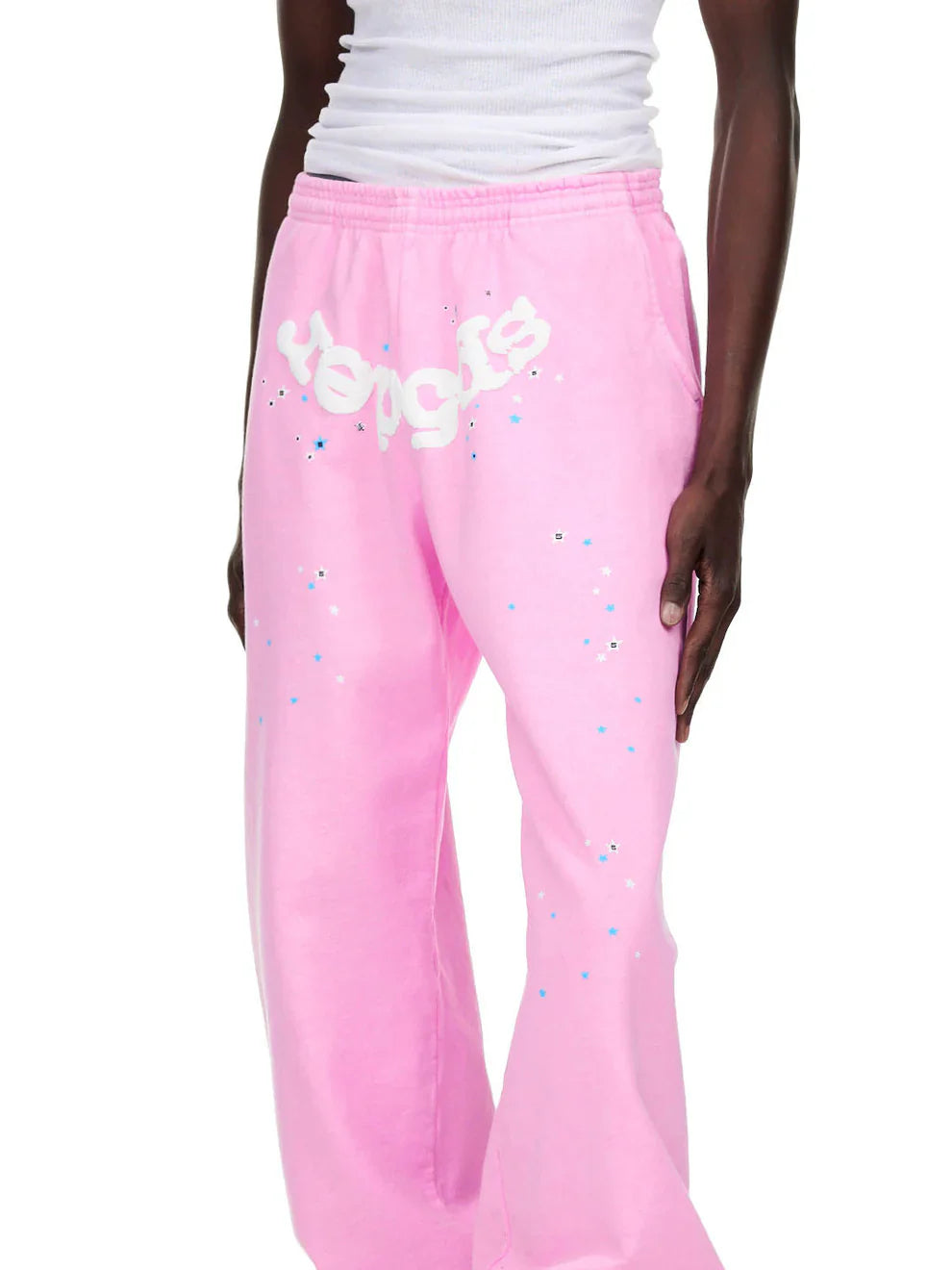 A person wears Sp5der Sweatpants – Unisex Light Pink Cotton Sweatpants featuring a front logo and star print, styled in a relaxed jogger fit, paired with a white sleeveless top for a casual streetwear look.