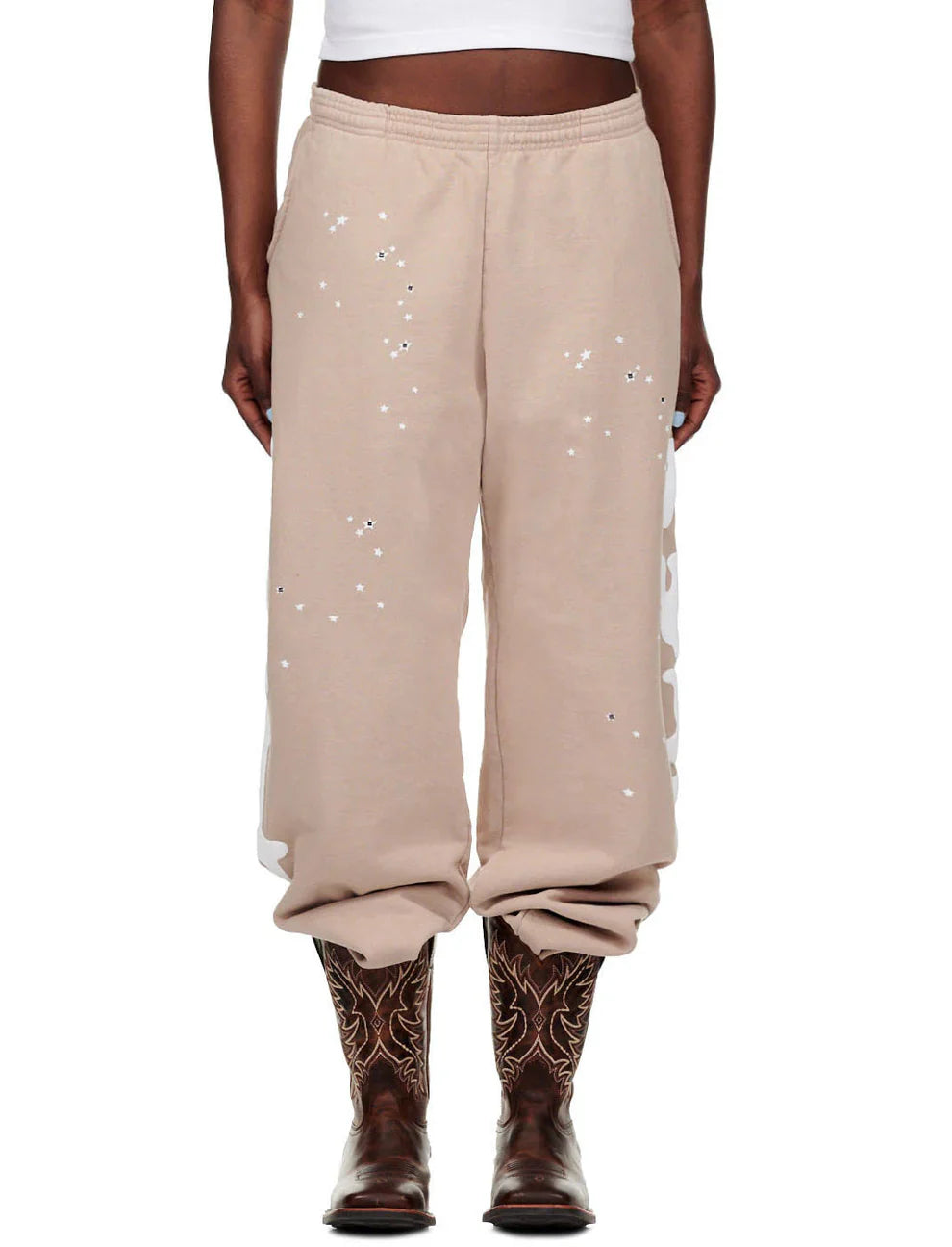 A person wearing 100% Cotton Sp5der Sweatpants for Men—beige streetwear jogger pants with side logo graphic and relaxed fit—paired with a white crop top and brown cowboy boots, stands against a plain background.