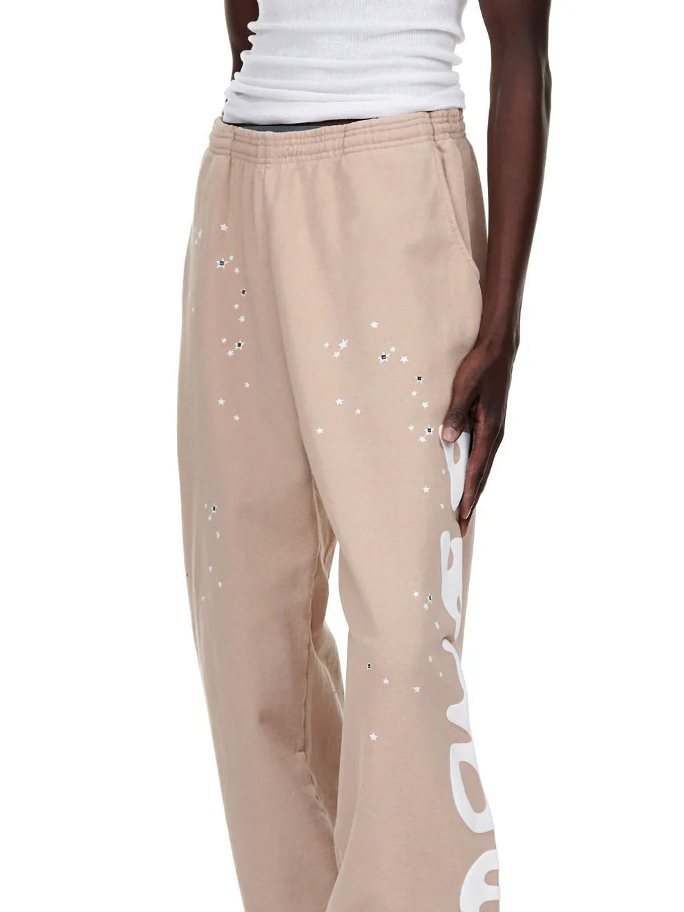 A person wears 100% Cotton Sp5der Sweatpants for Men—beige streetwear jogger pants with a side logo graphic and relaxed fit—paired with a white ribbed tank top, giving off laid-back cotton streetwear vibes.
