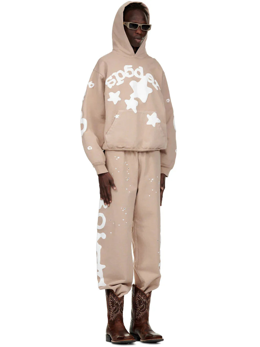 A person wears sunglasses, a beige hoodie with white designs, 100% Cotton Sp5der Sweatpants for Men – Beige Streetwear Jogger Pants with Side Logo Graphic, and brown embroidered cowboy boots, standing against a plain white background.