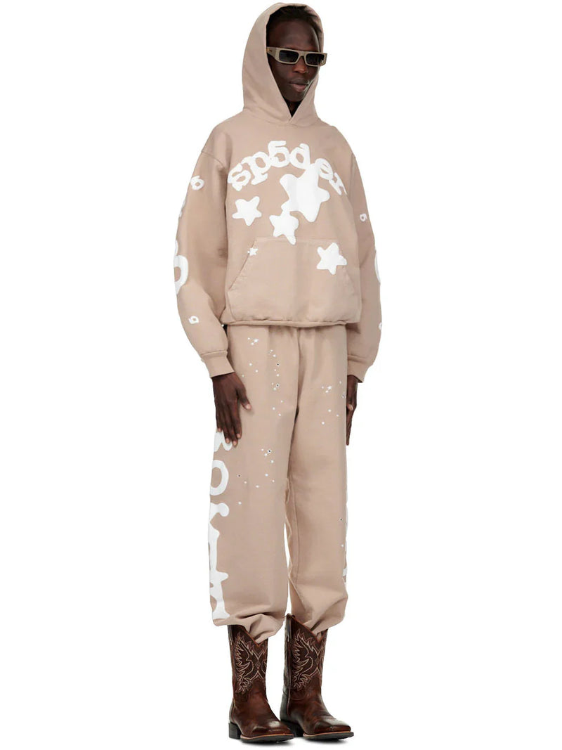 A person wears sunglasses, a beige hoodie with white designs, 100% Cotton Sp5der Sweatpants for Men – Beige Streetwear Jogger Pants with Side Logo Graphic, and brown embroidered cowboy boots, standing against a plain white background.