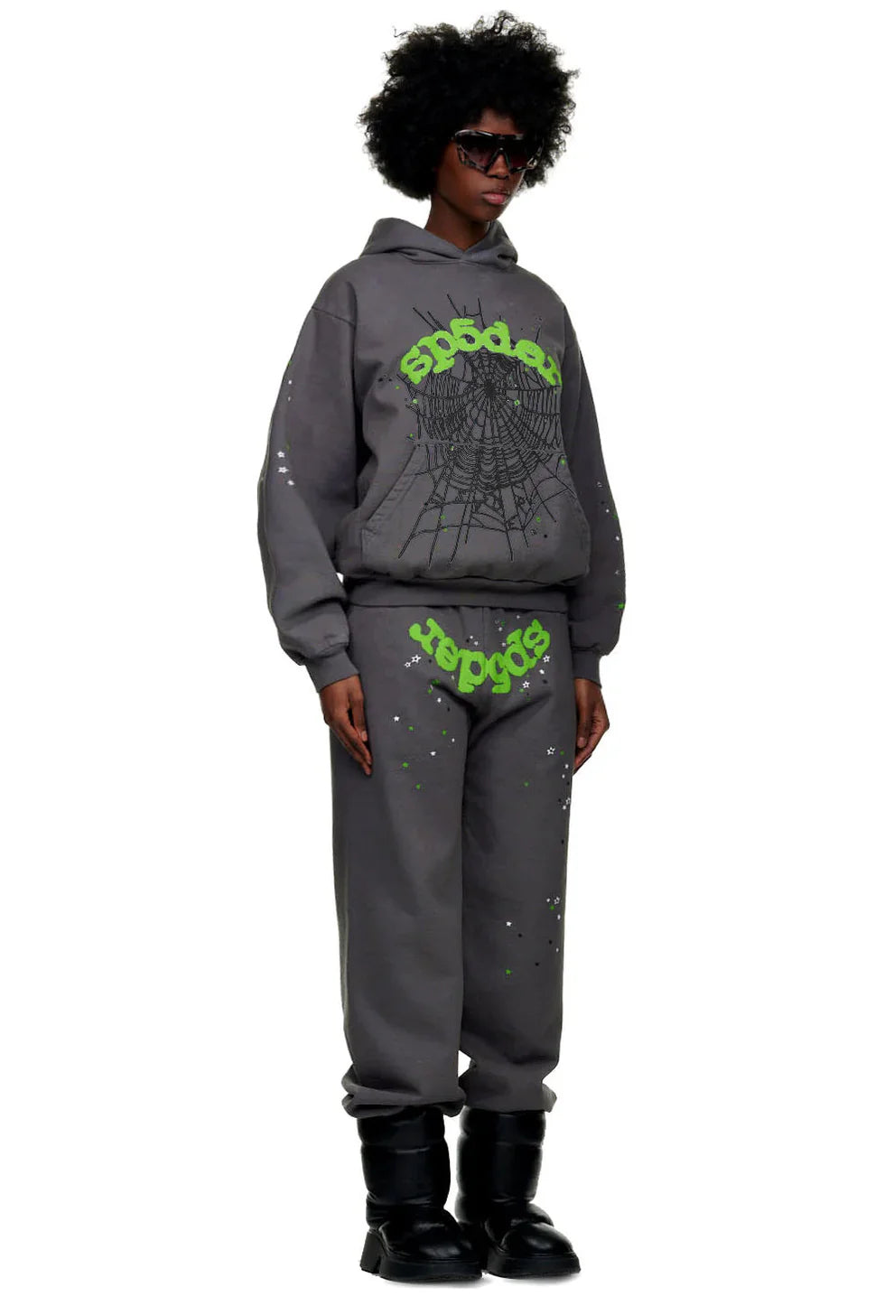 A man wears 100% cotton Sp5der sweatpants in dark grey with a green logo graphic and relaxed fit, paired with a matching hoodie, black sunglasses, and chunky boots, standing against a plain white background.