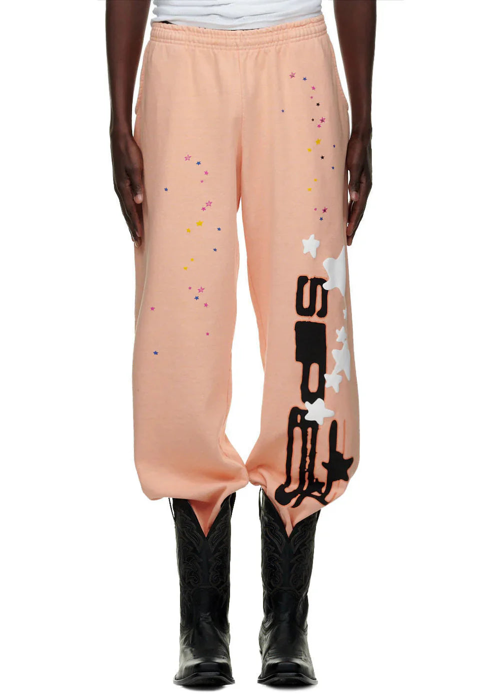 A person in 100% Cotton Sp5der Sweatpants—Unisex Peach Jogger Pants with Vertical Logo and Star Graphics, Relaxed Fit—styled with black cowboy boots, models streetwear against a plain background.