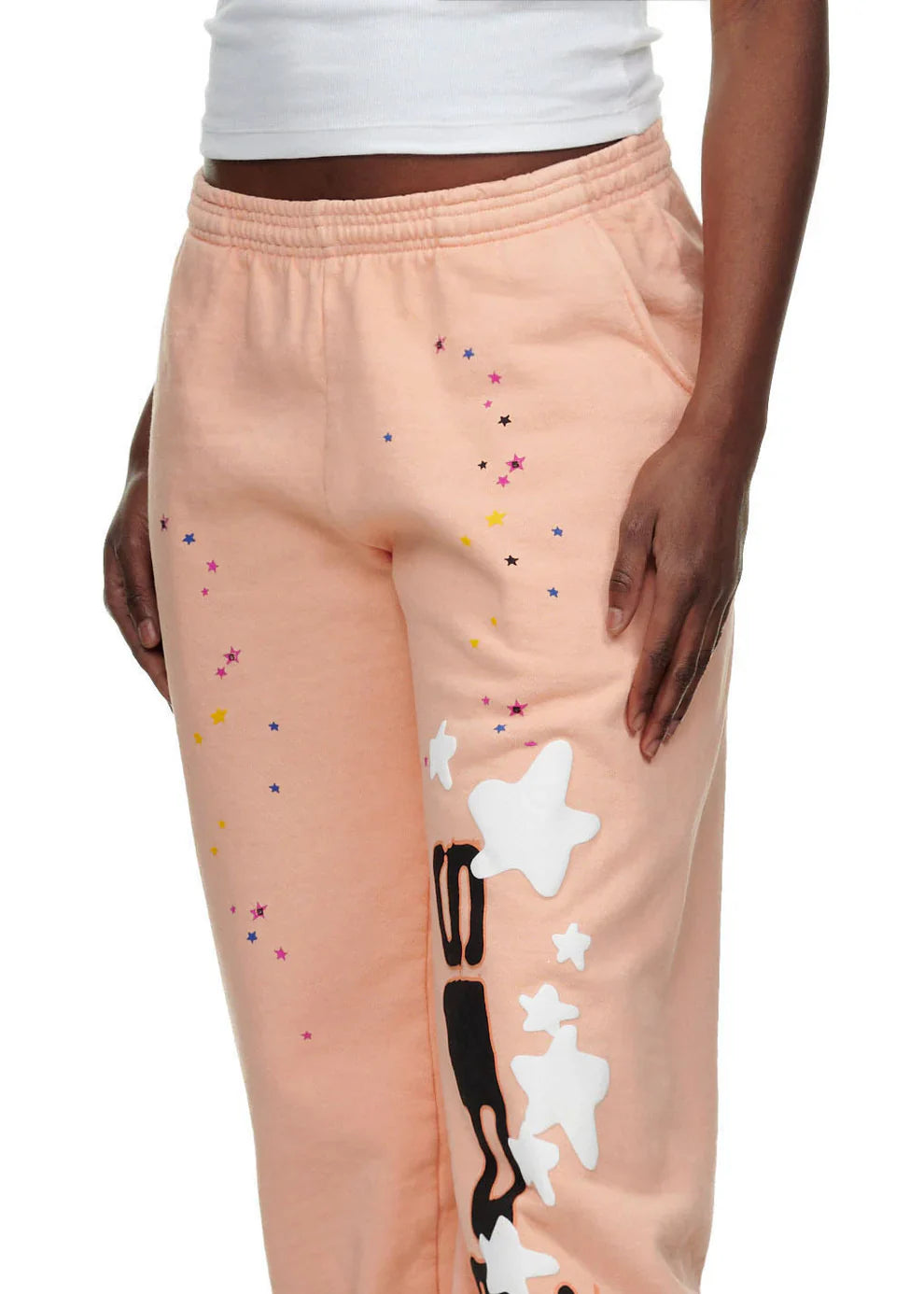 A person wearing 100% Cotton Sp5der Sweatpants – Unisex Peach Jogger Pants with vertical logo and star graphics, paired with a white cropped top; only their torso and upper legs are visible.