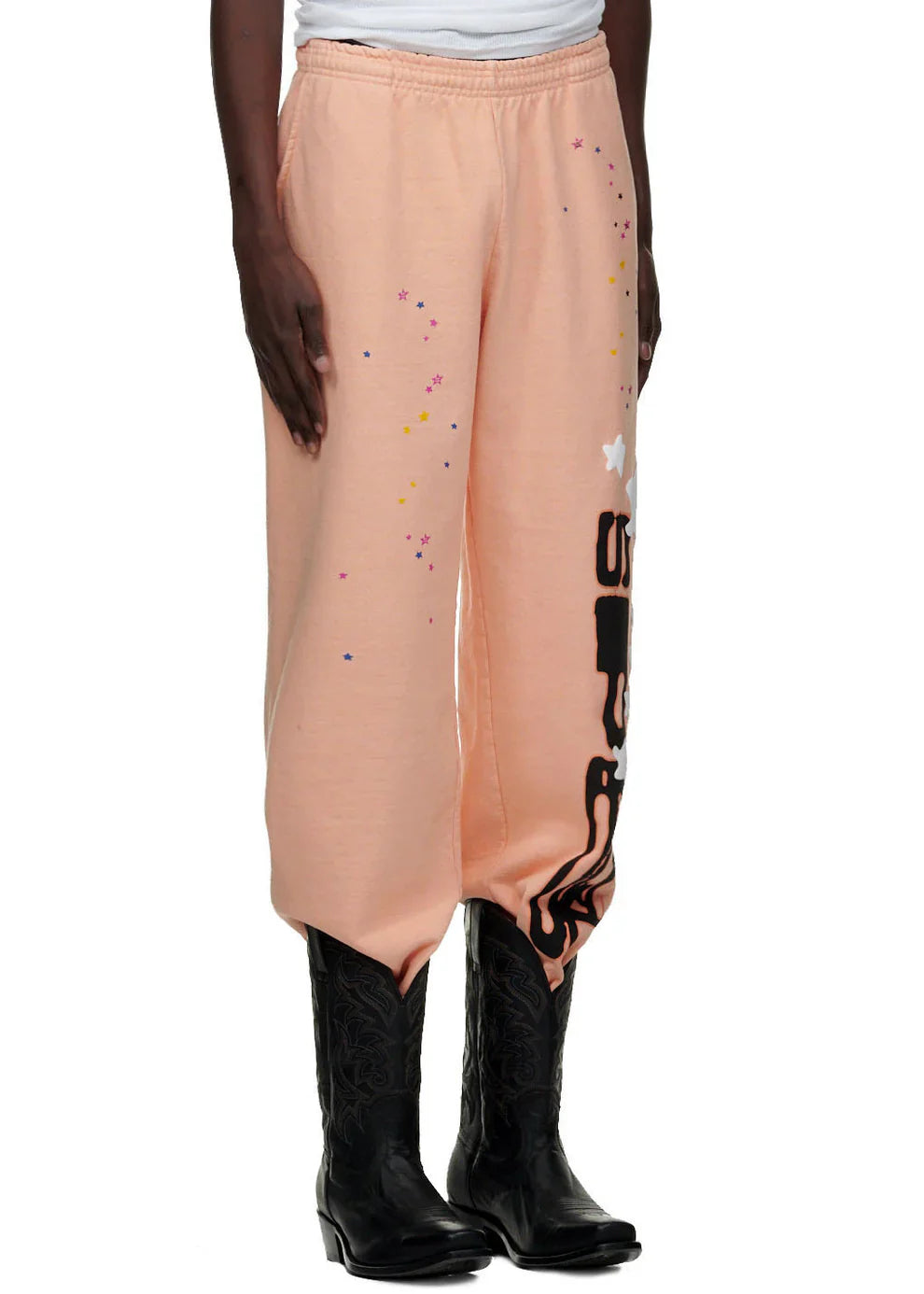 A person wears 100% cotton Sp5der Unisex Peach Jogger Pants featuring a vertical logo and star graphics, relaxed fit, paired with black cowboy boots. The upper body is mostly out of frame.