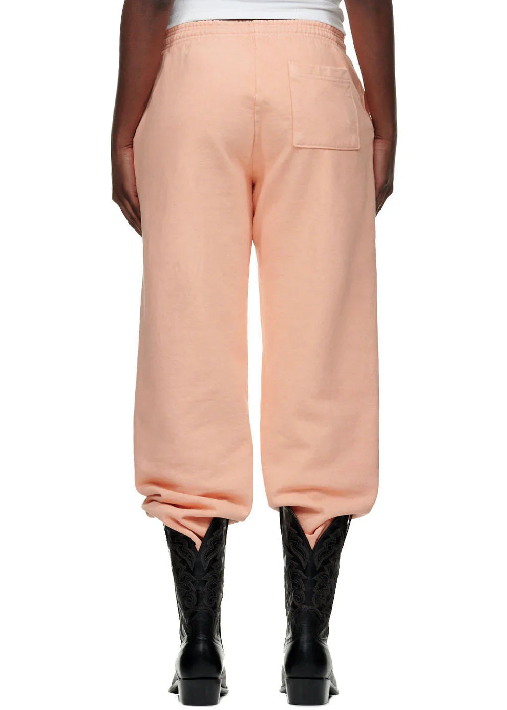 A person stands with their back to the camera wearing 100% Cotton Sp5der Peach Jogger Pants—unisex, relaxed fit with vertical logo and star graphics—paired with black textured cowboy boots and a white top for a streetwear vibe.
