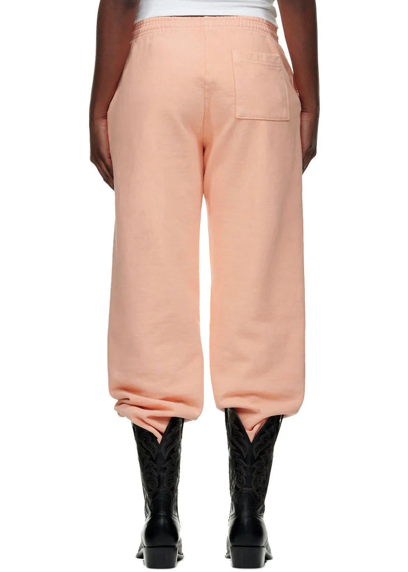 A person stands with their back to the camera wearing 100% Cotton Sp5der Peach Jogger Pants—unisex, relaxed fit with vertical logo and star graphics—paired with black textured cowboy boots and a white top for a streetwear vibe.