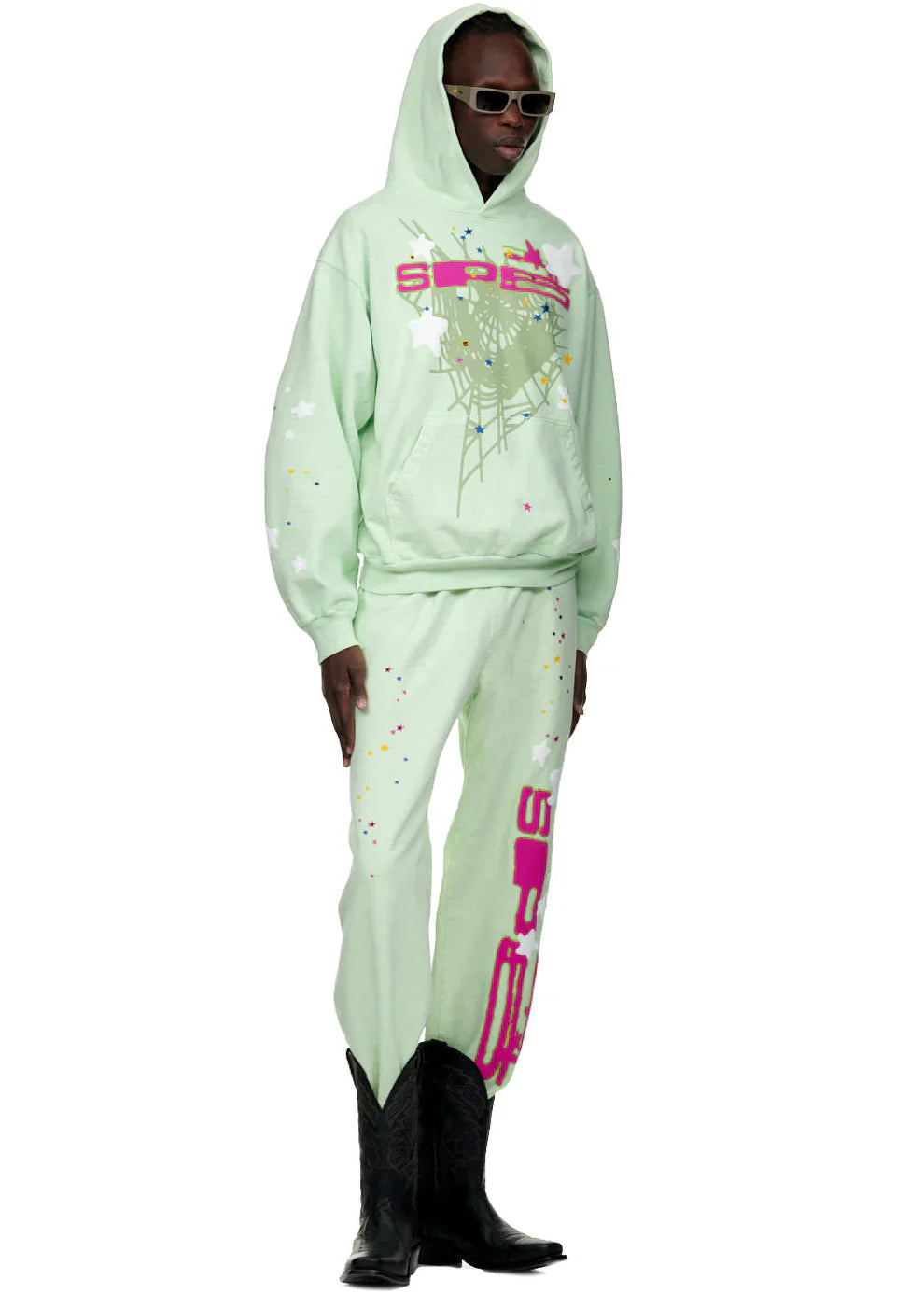 A person wears 100% cotton Sp5der unisex mint green jogger pants with vertical logo and star graphics, relaxed fit, styled boldly with a matching hoodie, black sunglasses, and cowboy boots against a white background.