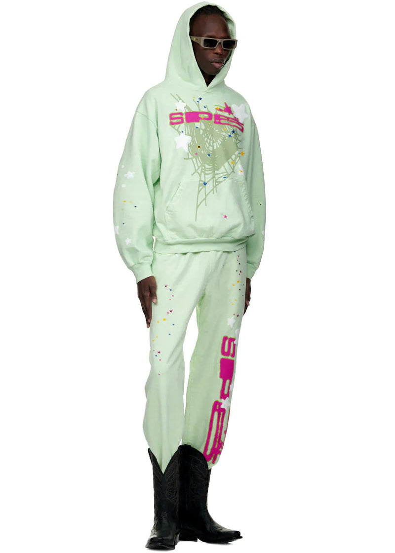 A person wears 100% cotton Sp5der unisex mint green jogger pants with vertical logo and star graphics, relaxed fit, styled boldly with a matching hoodie, black sunglasses, and cowboy boots against a white background.