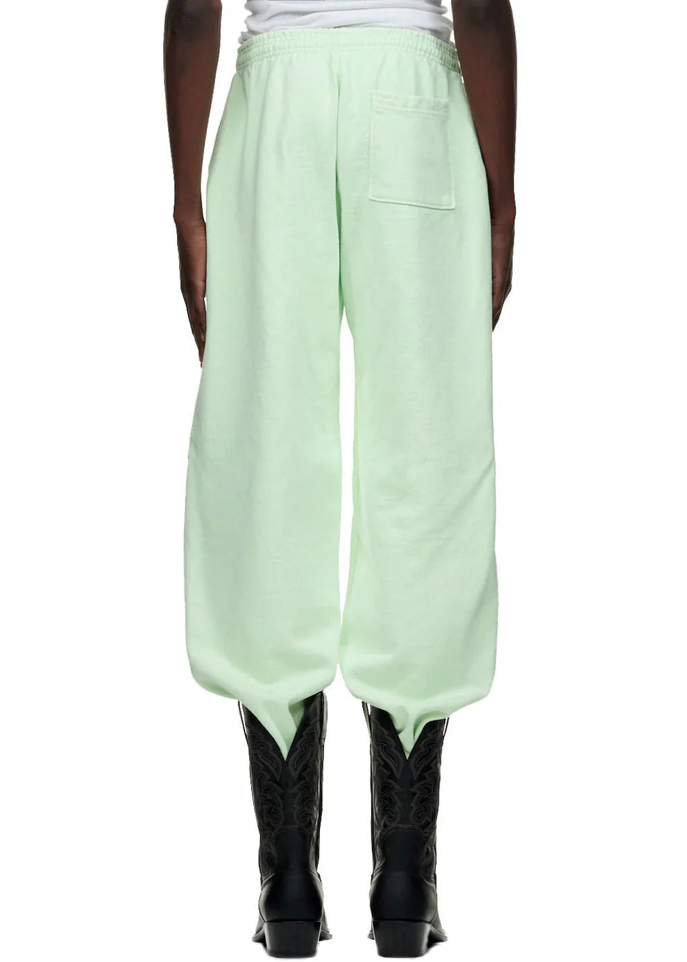 A person is seen from behind in 100% Cotton Sp5der Sweatpants – Unisex Mint Green Jogger Pants with vertical logo and star graphics, back pocket, paired with black embroidered cowboy boots and a white top against a plain white background.