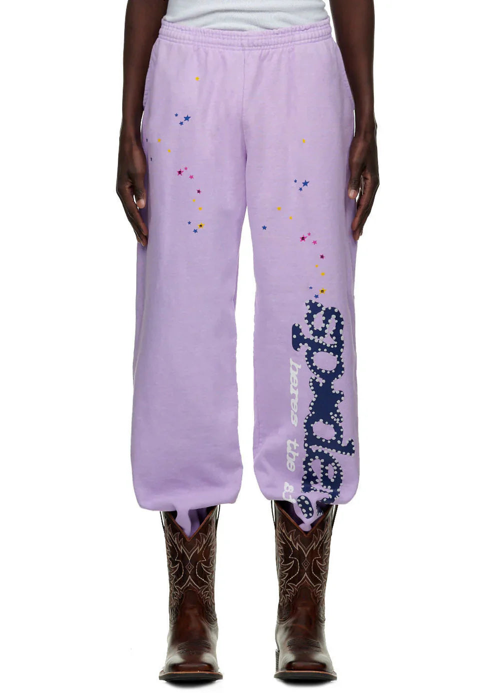 A person wears 100% Cotton Sp5der Unisex Lavender Jogger Pants with vertical logo and star details, styled with brown cowboy boots and a white shirt. The photo displays only the lower half of the body.