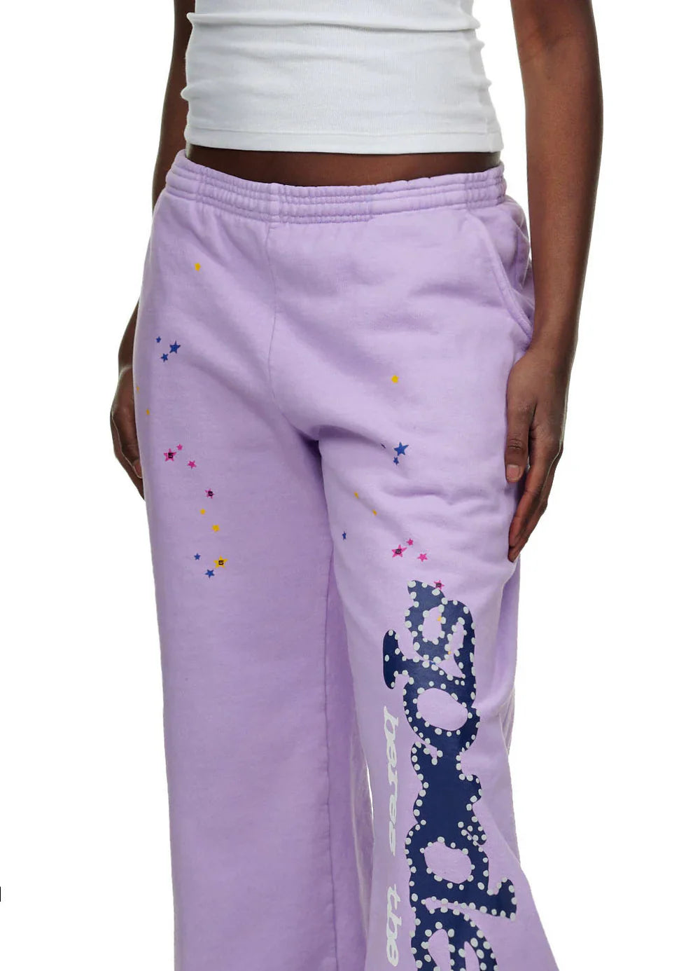 A person wears 100% Cotton Sp5der Unisex Lavender Jogger Pants with a vertical logo and star details, paired with a white sleeveless top. The streetwear look shows only the lower torso and part of one arm.