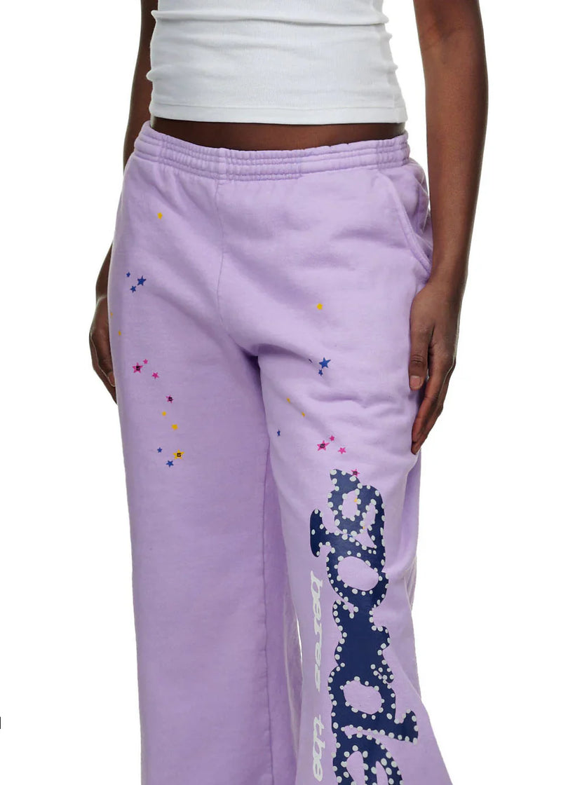A person wears 100% Cotton Sp5der Unisex Lavender Jogger Pants with a vertical logo and star details, paired with a white sleeveless top. The streetwear look shows only the lower torso and part of one arm.