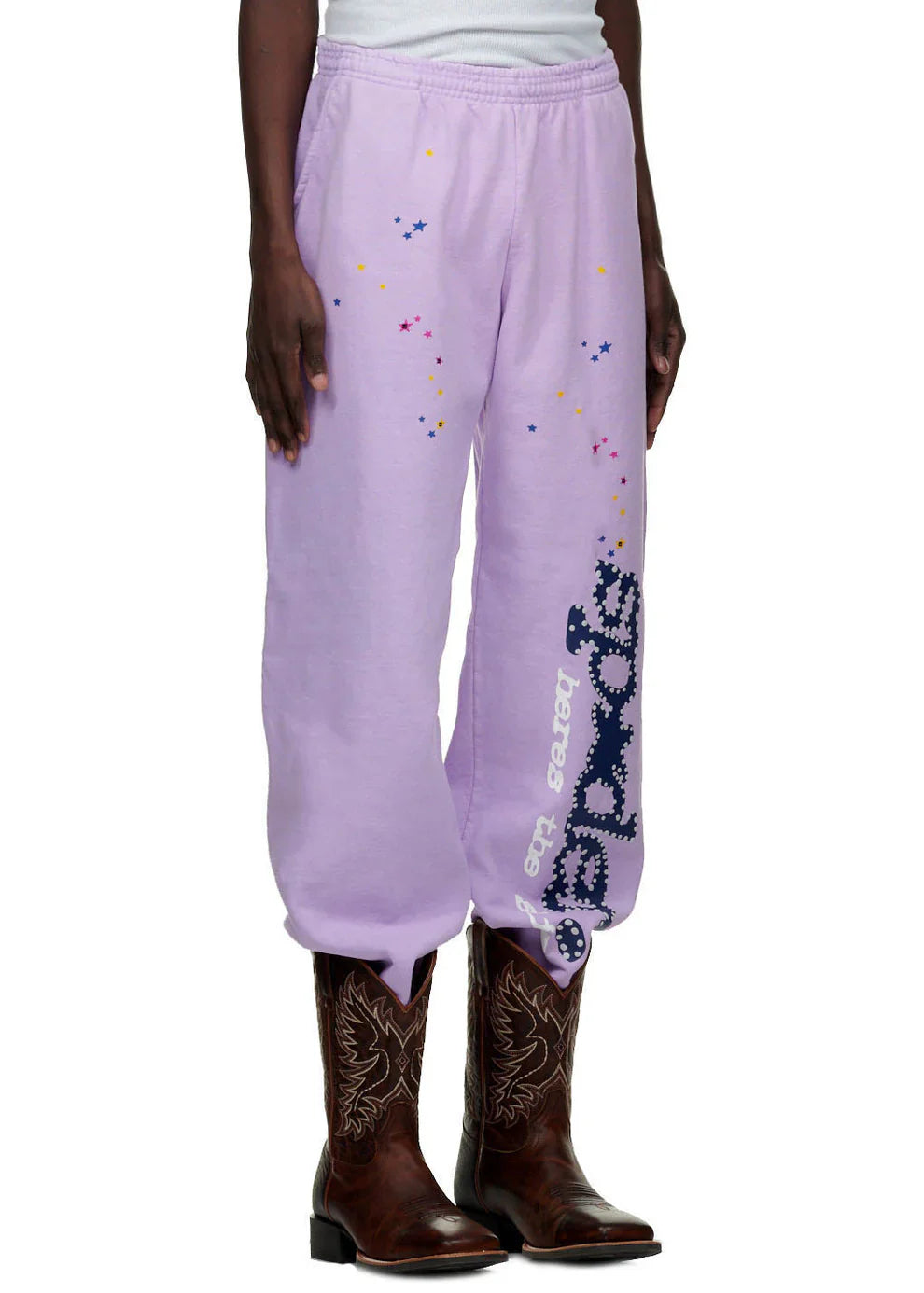 A person wears 100% Cotton Sp5der Sweatpants—Unisex Lavender Jogger Pants with vertical logo and star details, relaxed fit—paired with brown embroidered cowboy boots for a trendy streetwear look.