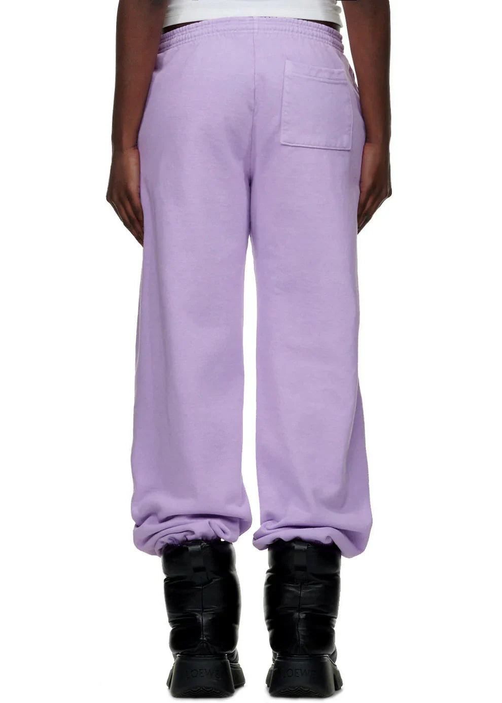 A person stands with their back to the camera, showing only lower back and legs in 100% cotton Sp5der Unisex Lavender Jogger Pants, featuring a relaxed fit, vertical logo, star details, gathered ankles, and a back pocket. Black boots complete the look.