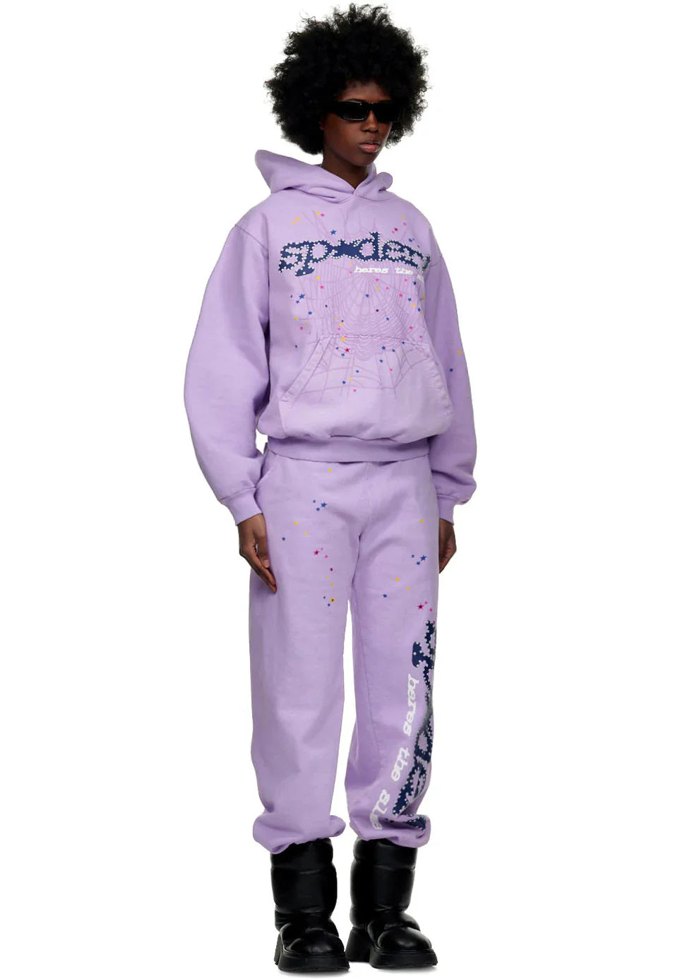 A person wears sunglasses, a lavender hoodie with a dark graphic and colorful dots, 100% Cotton Sp5der Sweatpants—Unisex Lavender Jogger Pants with vertical logo and star details—plus black boots, showcasing streetwear style on a white background.