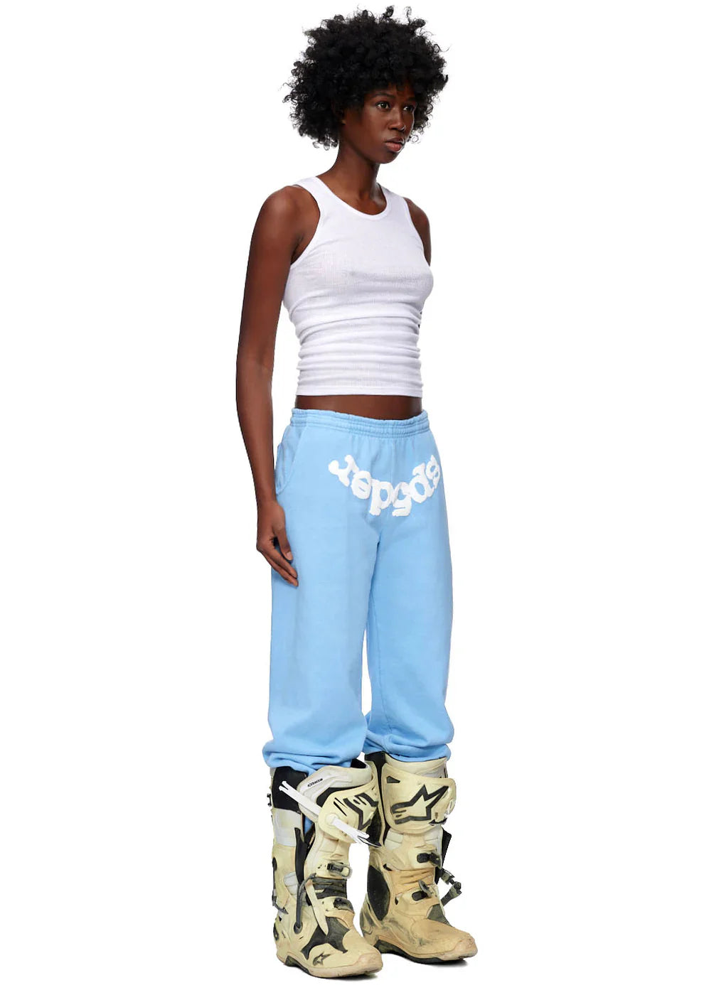 A person with a natural afro hairdo wears a fitted white tank top, Sp5der Unisex Light Blue Cotton Sweatpants with a front logo print, and large beige motocross boots, posing against a plain white background for a bold streetwear look.