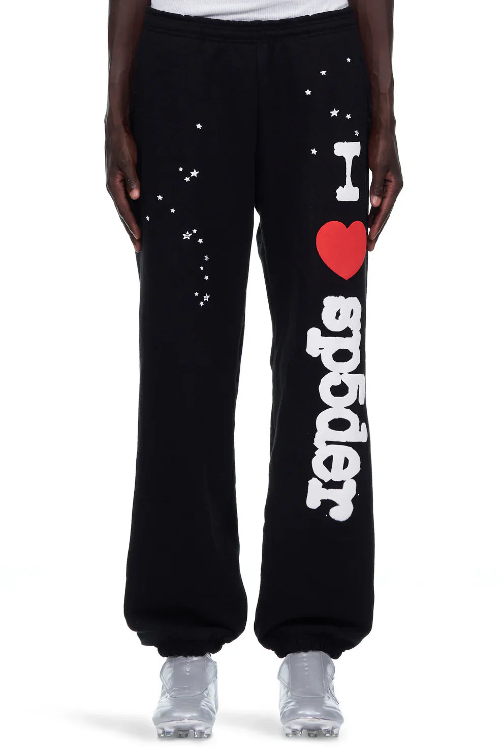 Photo shows a person from the waist down wearing unisex 100% cotton Sp5der Sweatpants—black jogger pants with a white “I Love Sp5der” graphic and small stars on the left leg—styled with white sneakers.