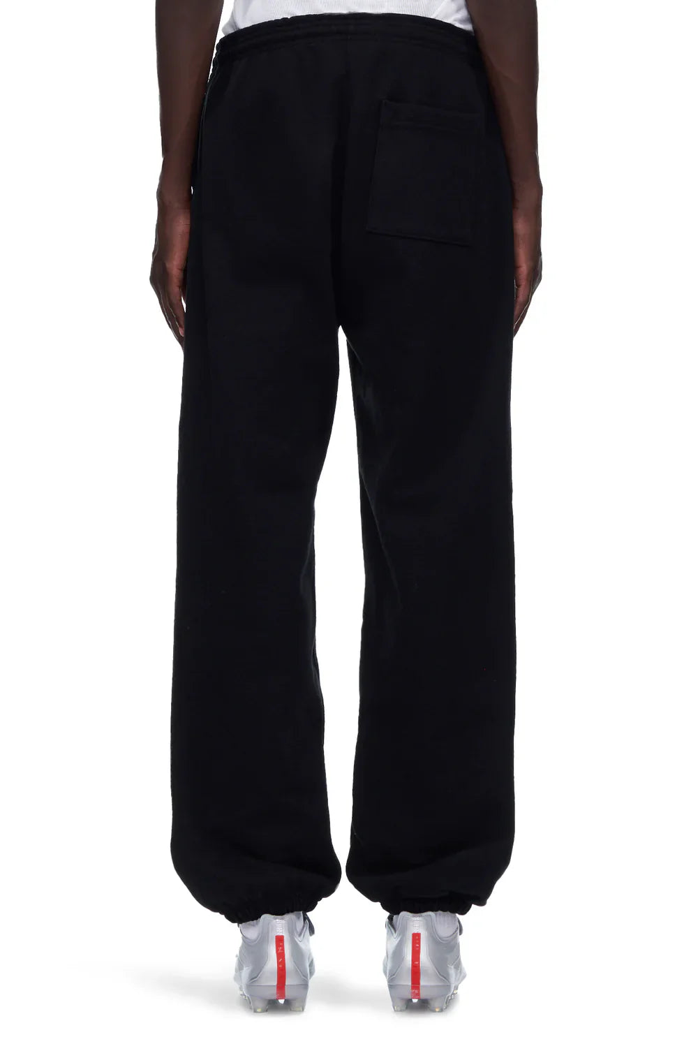 A person faces away wearing 100% Cotton Sp5der Sweatpants—Unisex Black Jogger Pants with "I Love Sp5der" graphic and a relaxed fit—paired with white sneakers featuring red accents against a plain white background.