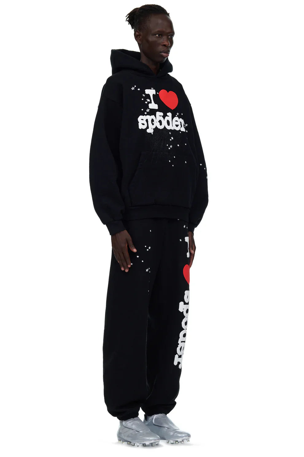 A person wears 100% Cotton Sp5der Sweatpants—unisex black jogger pants with an “I Love Sp5der” graphic and relaxed fit—styled with a matching black hoodie and silver athletic shoes for a bold streetwear look.