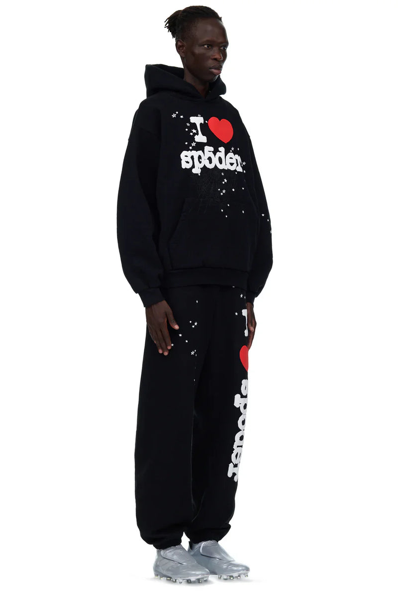 A person wears 100% Cotton Sp5der Sweatpants—unisex black jogger pants with an “I Love Sp5der” graphic and relaxed fit—styled with a matching black hoodie and silver athletic shoes for a bold streetwear look.