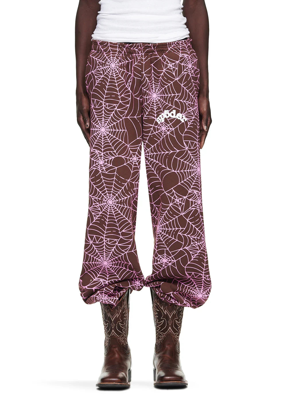 A person wears 100% Cotton Sp5der Sweatpants—Unisex Brown Jogger Pants with an all-over spider web print and relaxed fit—paired with a white shirt and cowboy boots for a stylish, unisex streetwear look. The pants bunch at the ankles.