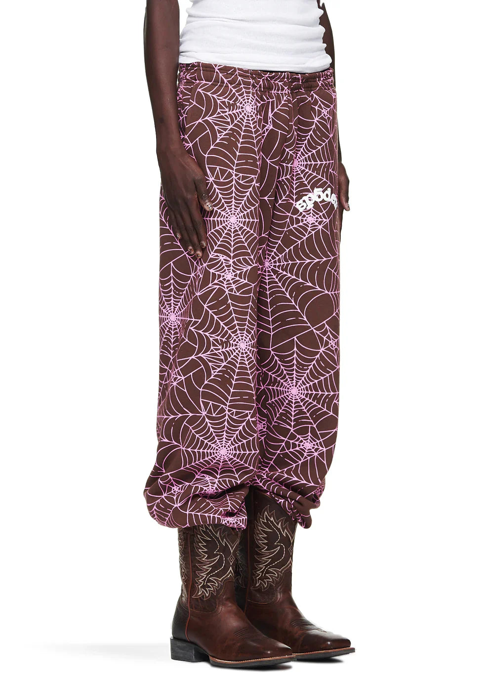 Someone in 100% Cotton Sp5der Sweatpants—Unisex Brown Jogger Pants with All-Over Spider Web Print and a relaxed fit—wears a white top and brown cowboy boots against a white background for bold streetwear style.