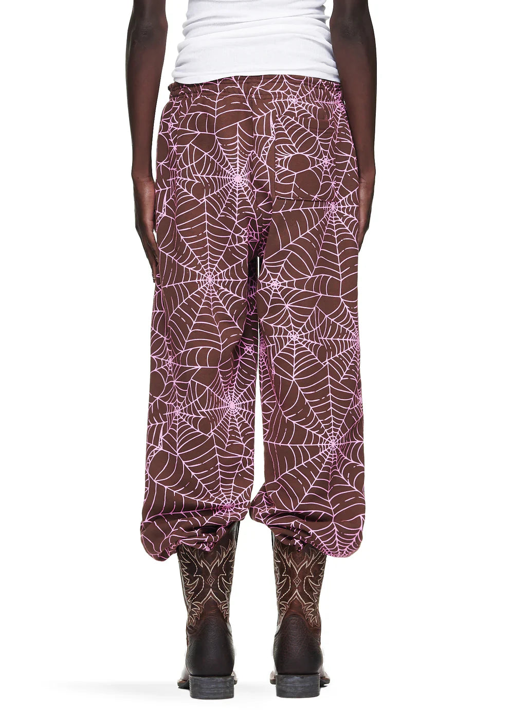 Someone seen from behind wears 100% Cotton Sp5der Sweatpants—Unisex Brown Jogger Pants with All-Over Spider Web Print, relaxed fit, paired with a white top and brown boots. The loose joggers are gathered at the ankles.