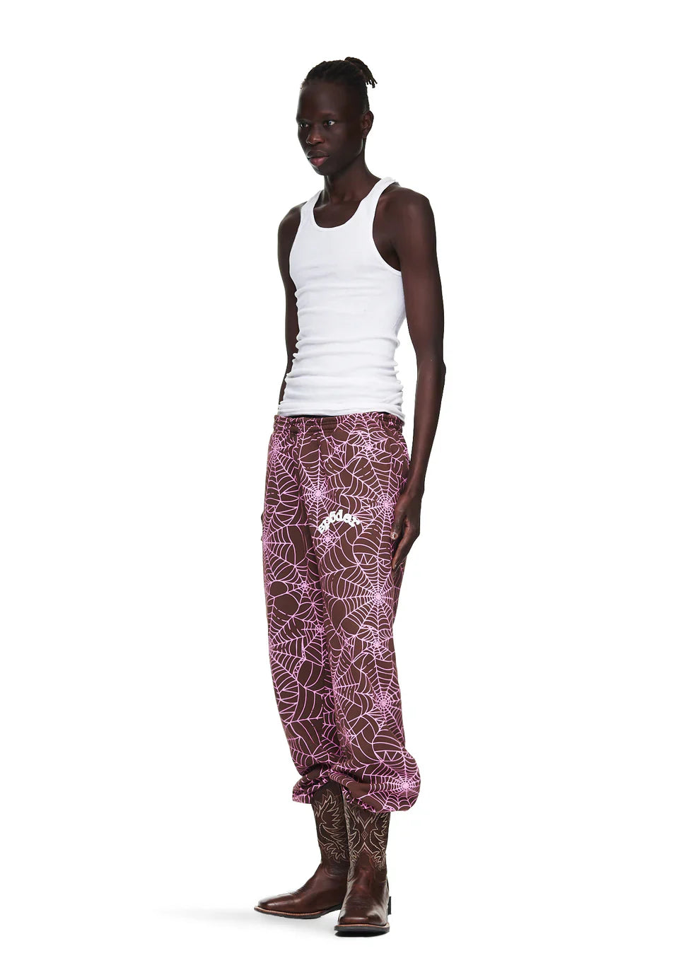 A person wearing a white tank top, 100% Cotton Sp5der Unisex Brown Jogger Pants with all-over spider web print (relaxed fit), and cowboy boots stands against a white background.