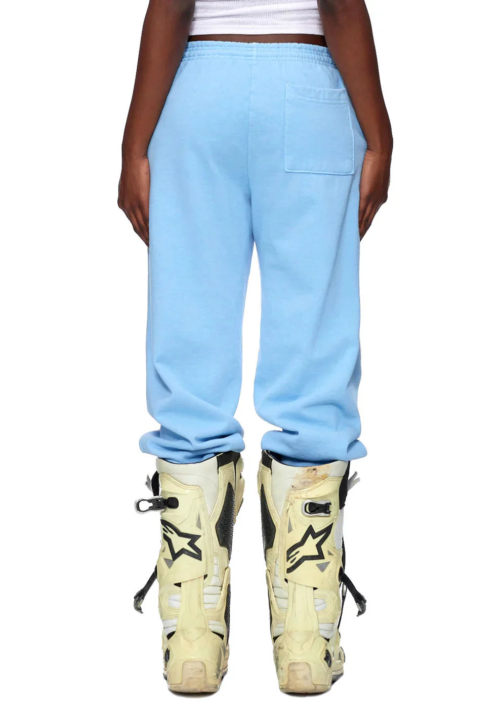 A person shown from the back in Sp5der Sweatpants—unisex light blue cotton sweatpants with front logo print—paired with a white ribbed tank and tall white motocross boots featuring black stars, set against a white background for bold streetwear flair.
