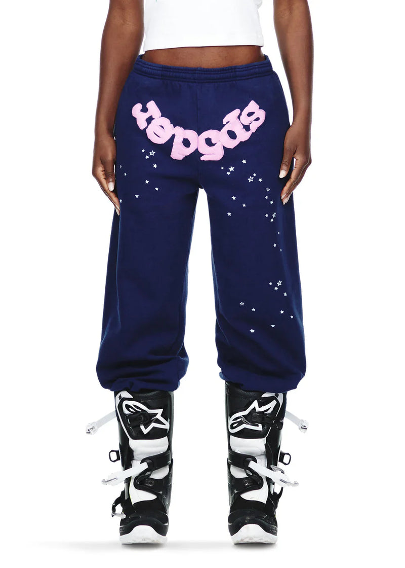 A person wears 100% Cotton Sp5der Sweatpants—unisex navy blue jogger pants with a pink logo and star graphics, relaxed fit—paired with black and white motocross boots. Their torso is cropped out of the image.