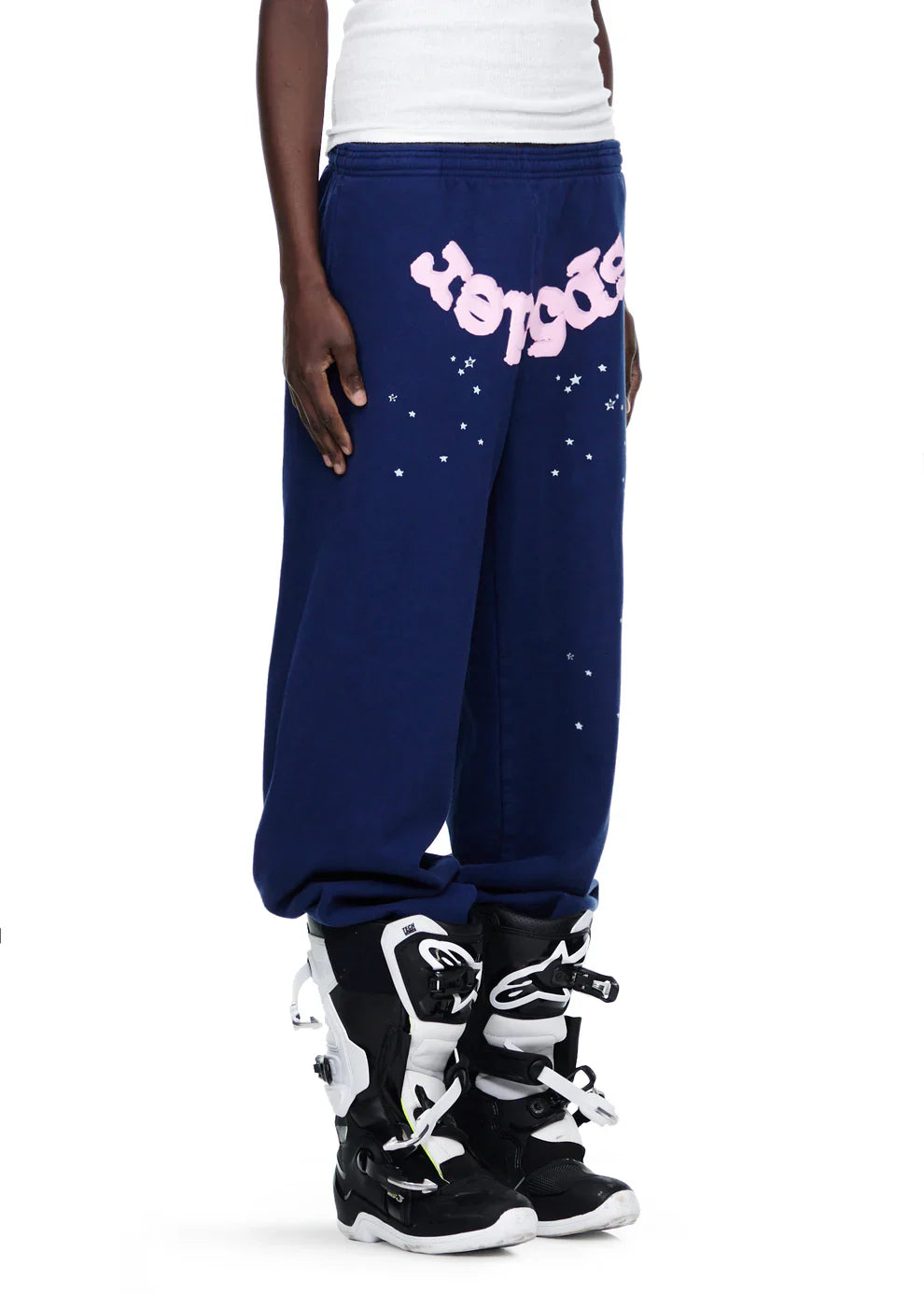 A person wears 100% cotton Sp5der unisex navy blue jogger pants with a pink logo and star graphics, styled with black and white motocross boots and a white top for a bold streetwear look.