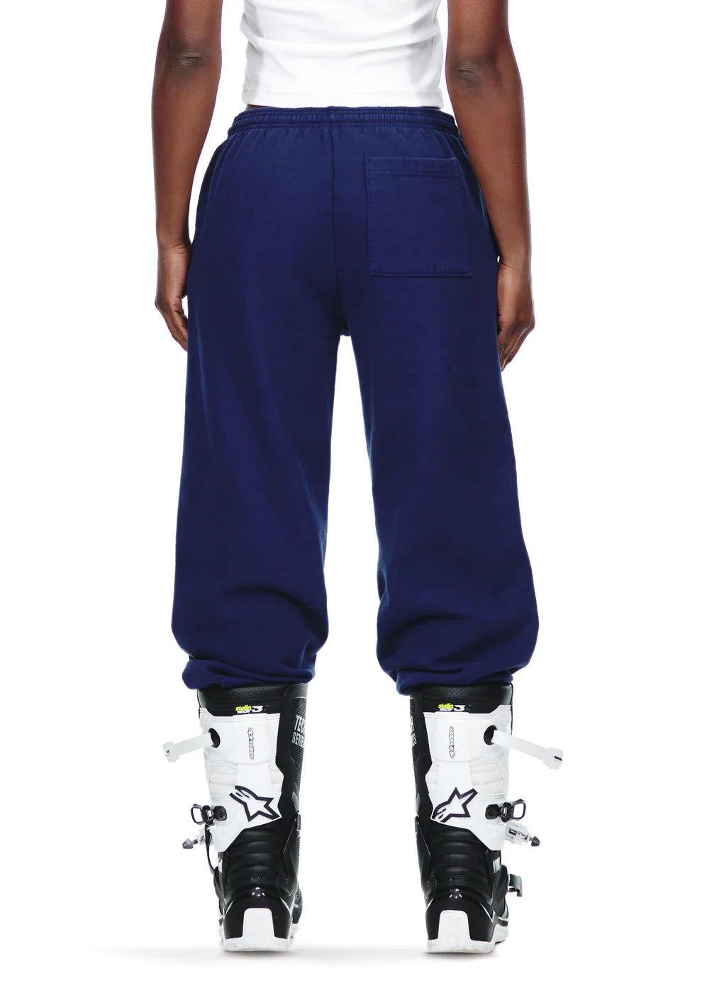 A person wearing a white shirt, 100% Cotton Sp5der Sweatpants – Unisex Navy Blue Jogger Pants with Pink Logo and Star Graphics, and black and white ski boots stands facing away, hands relaxed by their sides.