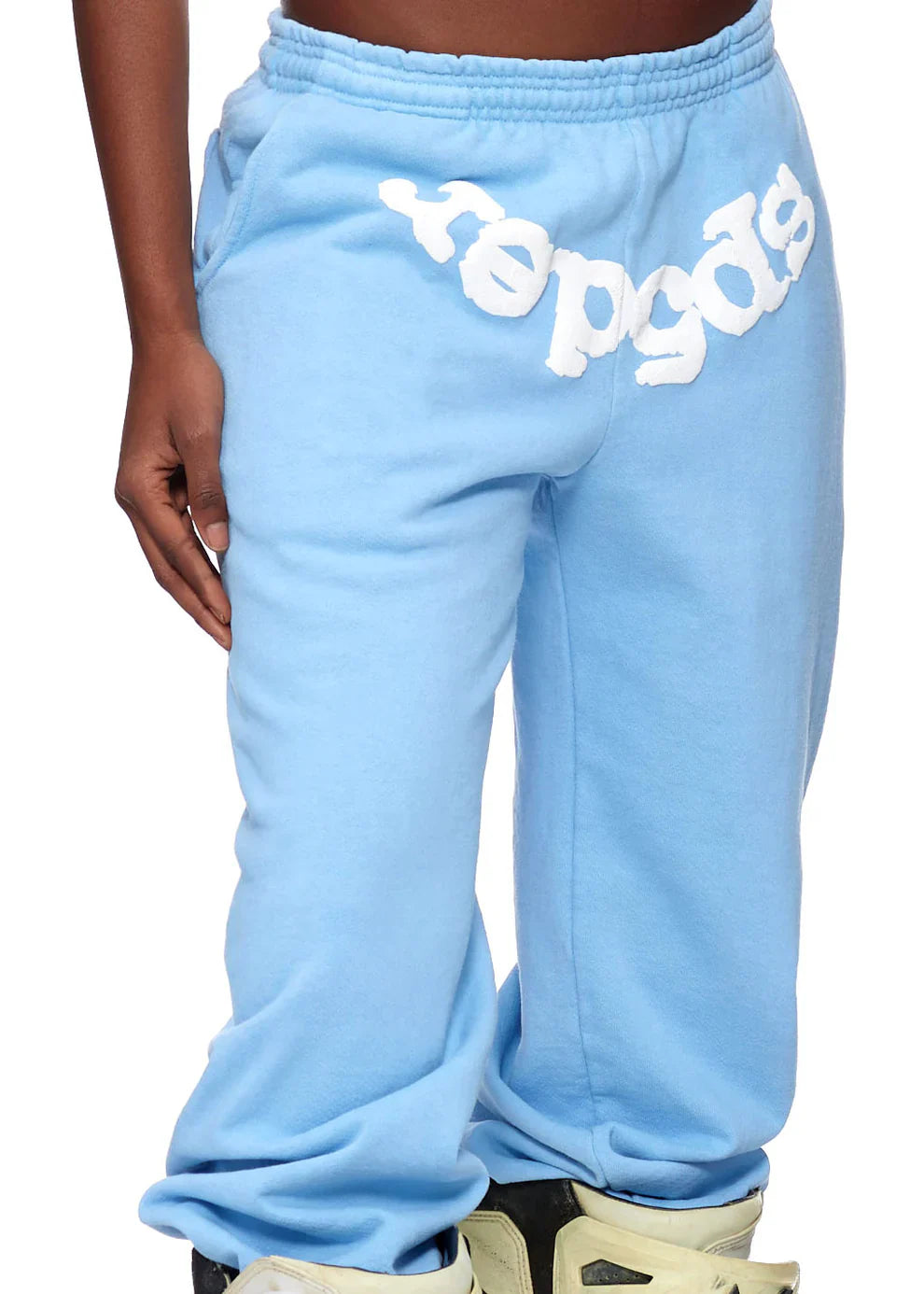 Someone showcases modern streetwear in Sp5der Sweatpants – Unisex Light Blue Cotton Sweatpants with Front Logo Print, paired with off-white sneakers. Only the lower torso and legs are shown, emphasizing the textured front logo print.