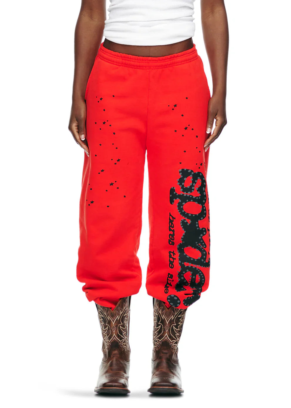 Someone wearing 100% Cotton Sp5der Sweatpants—Unisex Red Jogger Pants with black logo and star graphics, relaxed fit—with a white tank top and brown cowboy boots, showcases bold streetwear style against a white background.