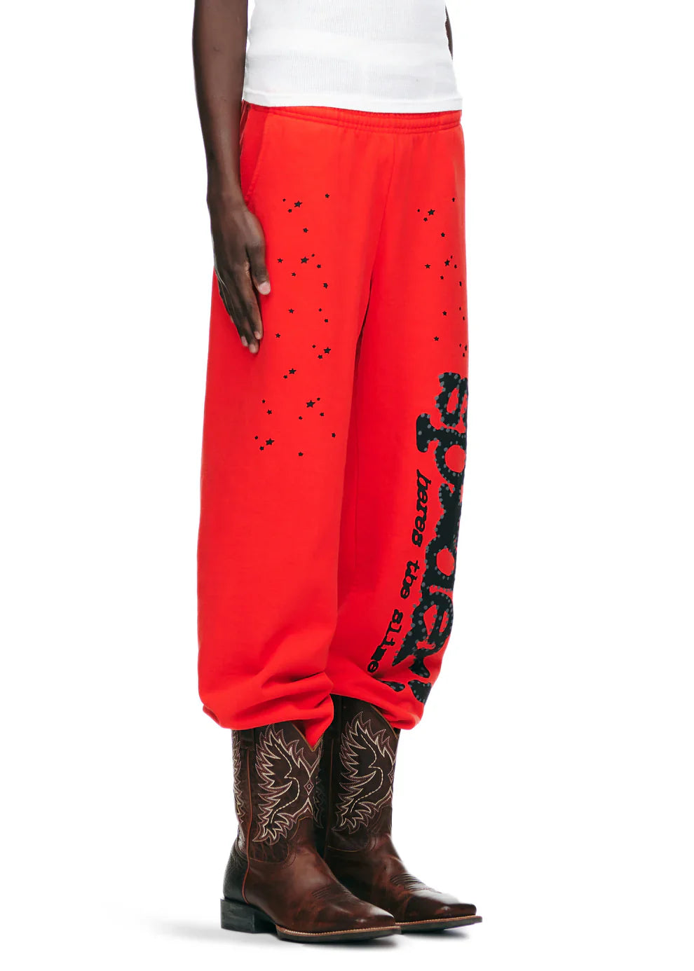 A person in 100% Cotton Sp5der Unisex Red Jogger Pants with black logo and star graphics, styled with a white top and cowboy boots, poses against a plain background—highlighting vibrant streetwear style.