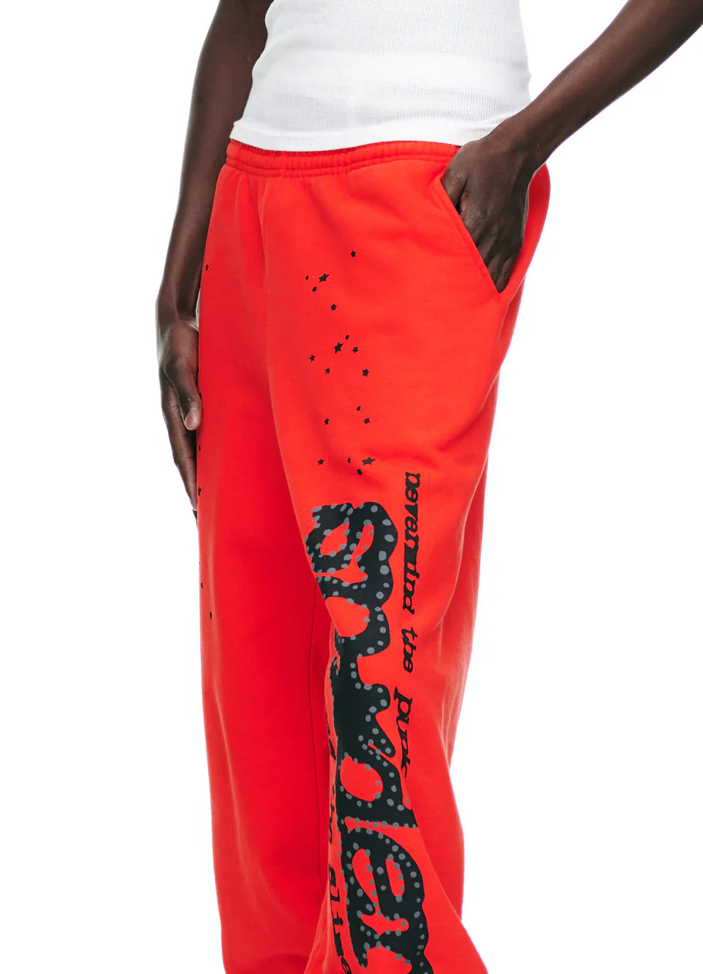 A person in 100% Cotton Sp5der Sweatpants—unisex red jogger pants with black logo and star graphics, relaxed fit—styled with a white sleeveless top, channels streetwear vibes, one hand casually tucked in their pocket.