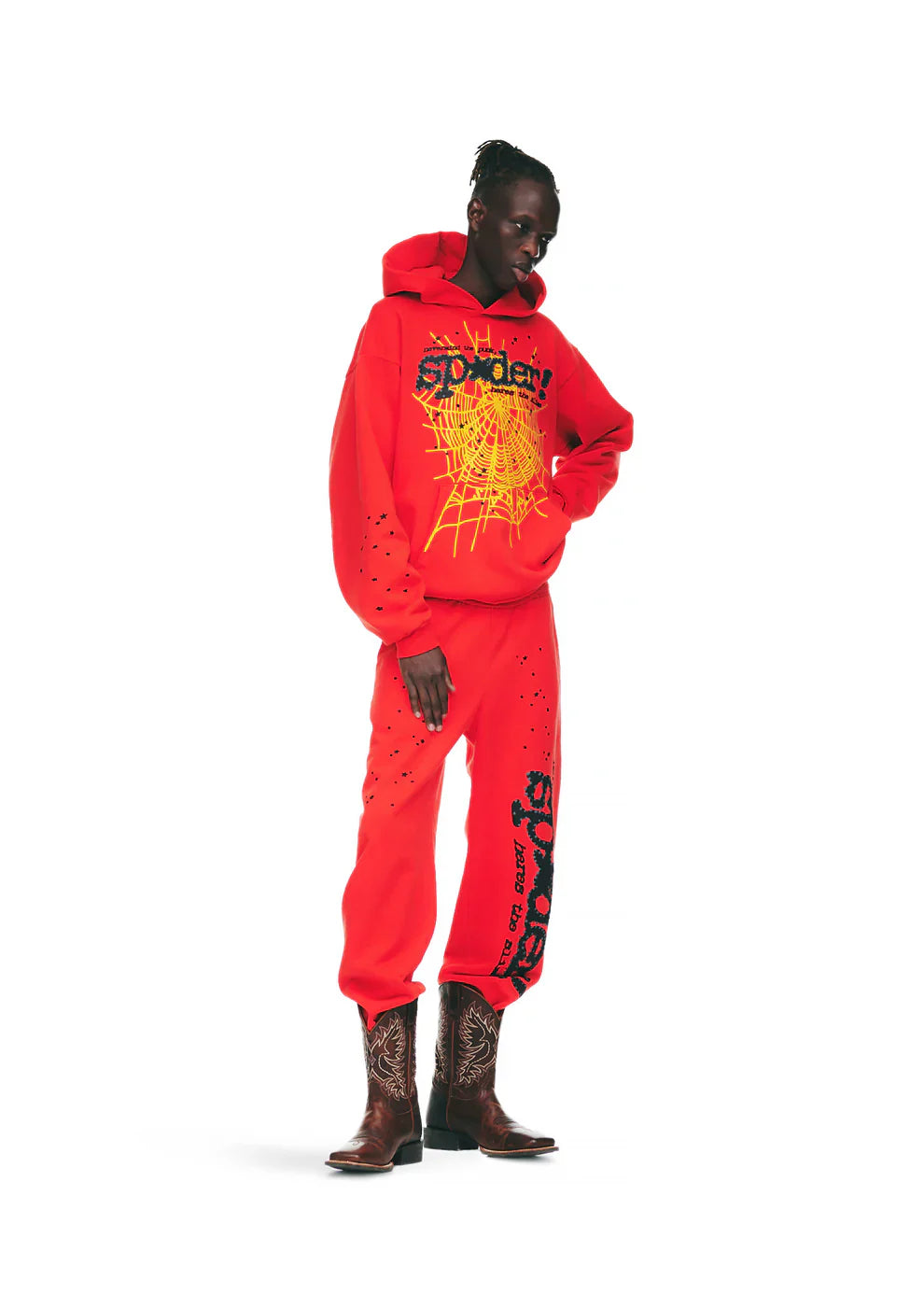 A person stands against a plain white background wearing 100% Cotton Sp5der Sweatpants—unisex red jogger pants with black logo and star graphics, relaxed fit—paired with a bright red hoodie and brown cowboy boots.