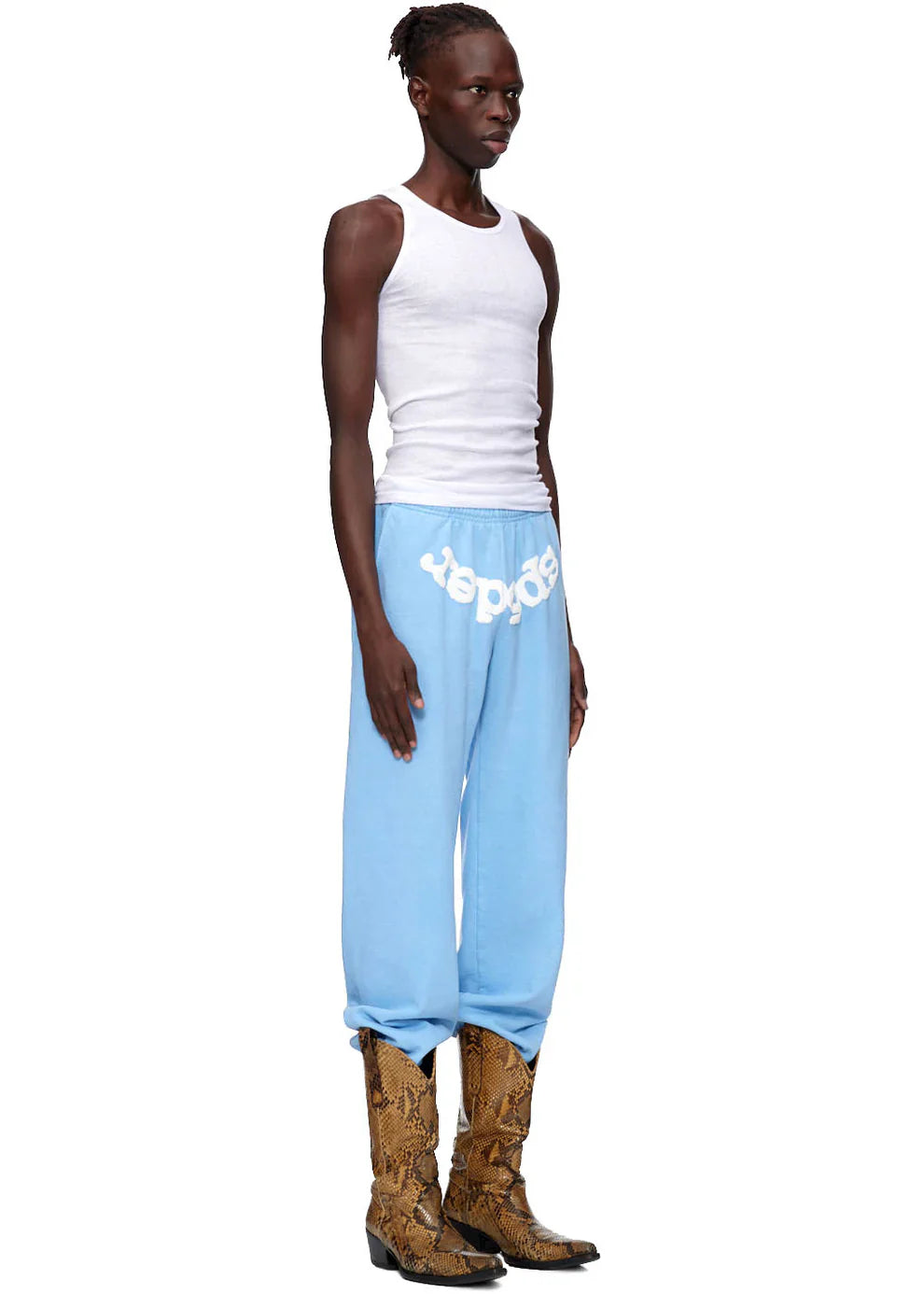 Wearing a white tank top, Sp5der Unisex Light Blue Cotton Sweatpants with front logo print, and tan snakeskin cowboy boots, a person faces left against a plain white background for a sleek streetwear-inspired look.