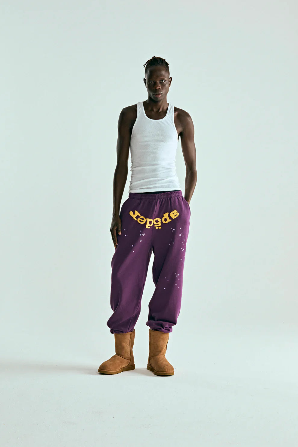 Against a plain background, a person wears white tank top, tan boots, and 100% Cotton Sp5der Sweatpants—Unisex Purple Jogger Pants with yellow logo and star graphics, featuring a relaxed fit. Their hands are in their pockets.