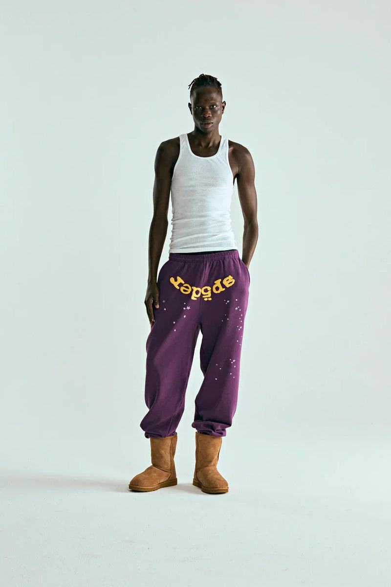 Against a plain background, a person wears white tank top, tan boots, and 100% Cotton Sp5der Sweatpants—Unisex Purple Jogger Pants with yellow logo and star graphics, featuring a relaxed fit. Their hands are in their pockets.