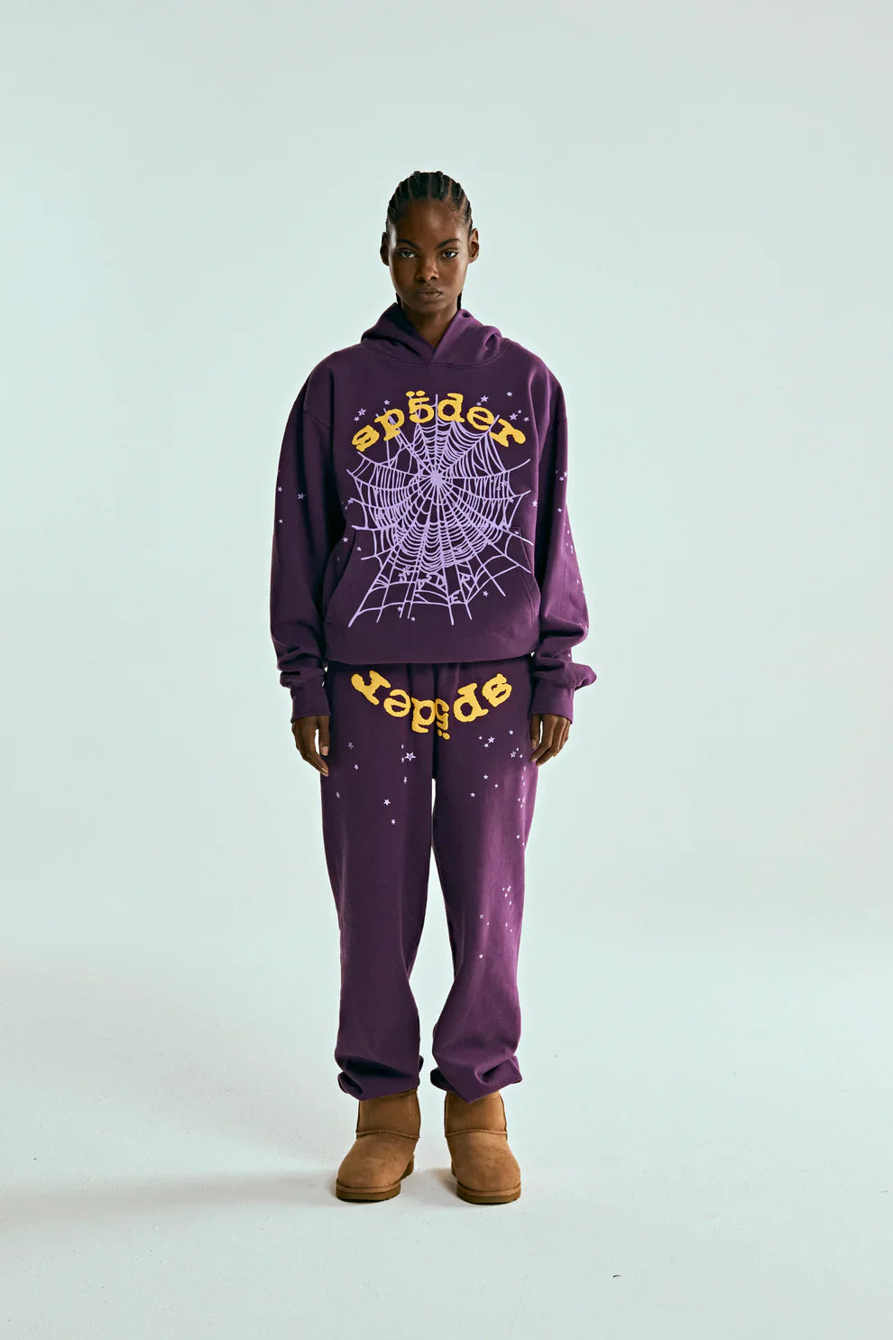 A person stands against a plain background in unisex streetwear—a purple hoodie and 100% Cotton Sp5der Sweatpants with a yellow logo and star graphics, relaxed fit—paired with tan boots.