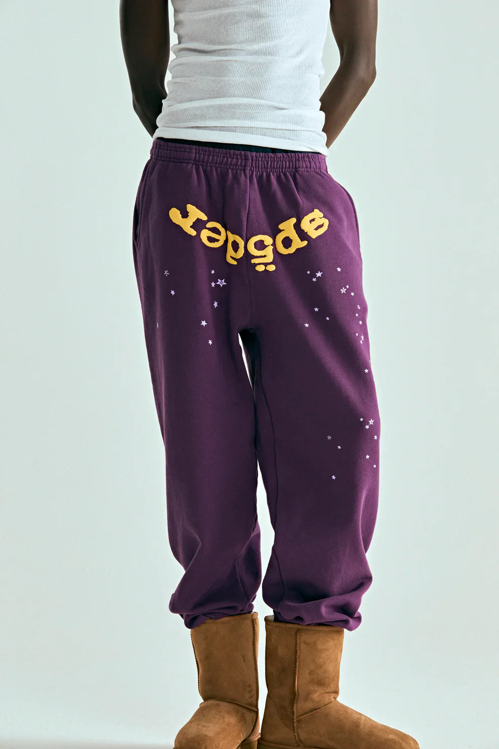 A person wearing a white tank top, tan boots, and 100% Cotton Sp5der Sweatpants—Unisex Purple Jogger Pants with a yellow logo and star graphics—stands against a plain background.