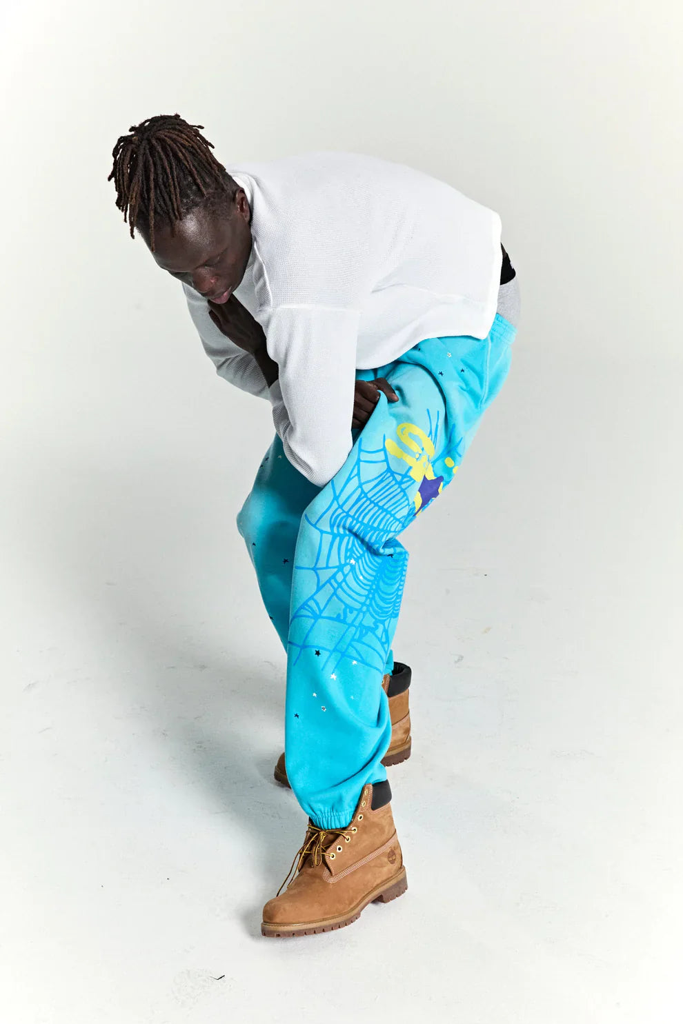 A person in a white sweater, 100% Cotton Sp5der Sweatpants—Unisex Blue Jogger Pants with spider web and star details—plus tan boots, bends down to adjust their pant leg in a white studio setting.