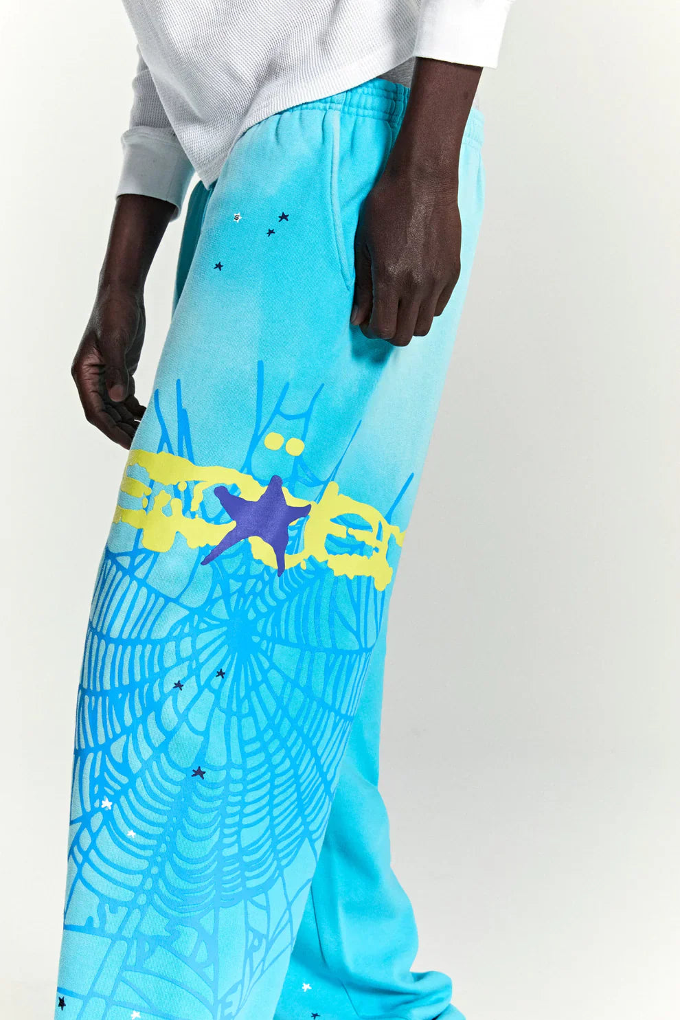 A person sports 100% Cotton Sp5der Sweatpants—unisex blue jogger pants with a spider web graphic and star details, relaxed fit—paired with a white long-sleeve shirt for a standout streetwear look.