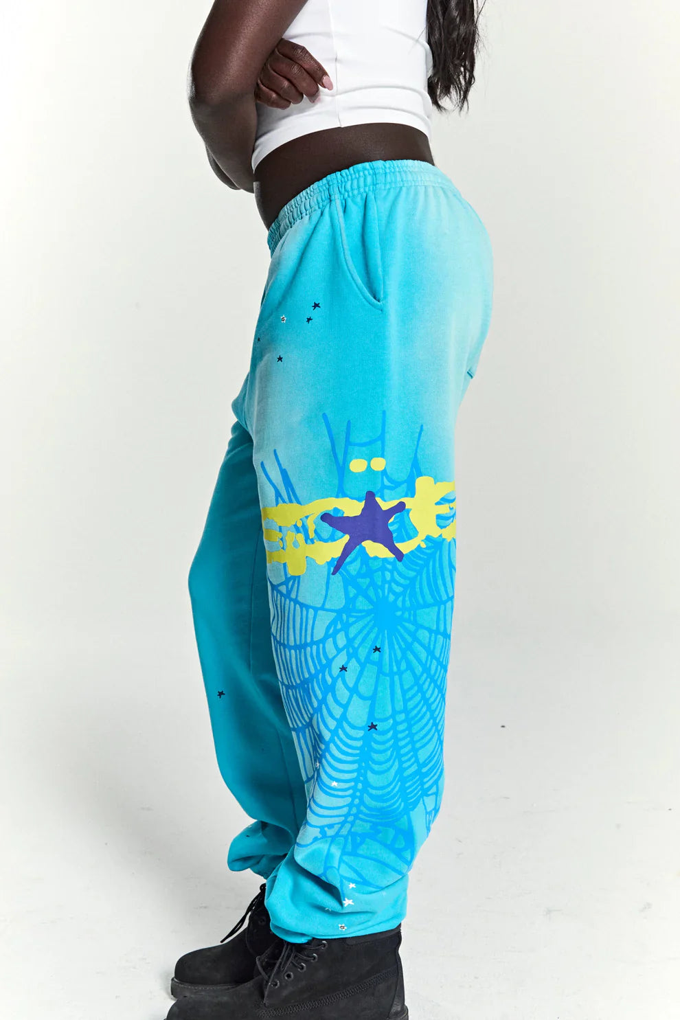 A person in a white crop top and black boots stands with arms crossed, wearing 100% Cotton Sp5der Sweatpants—Unisex Blue Jogger Pants with a spider web graphic and star details—showcasing bold streetwear against a plain white background.