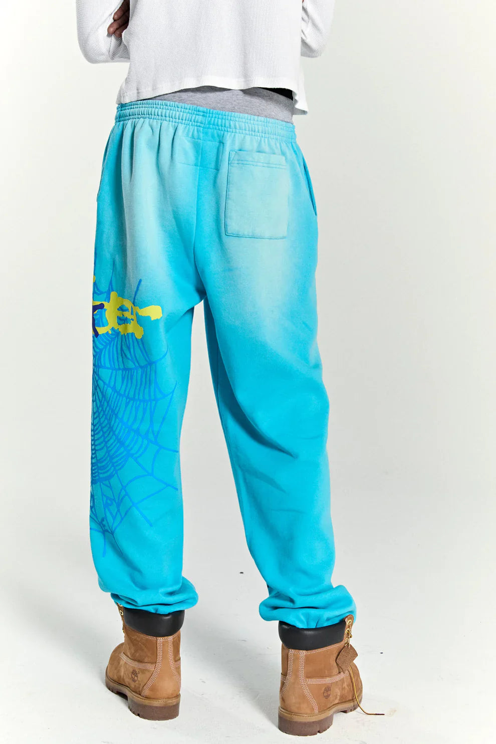 Person stands with their back to the camera in 100% cotton Sp5der sweatpants—unisex blue joggers with spider web and star graphics, relaxed fit—paired with a white long-sleeve shirt, grey waistband, and tan work boots.