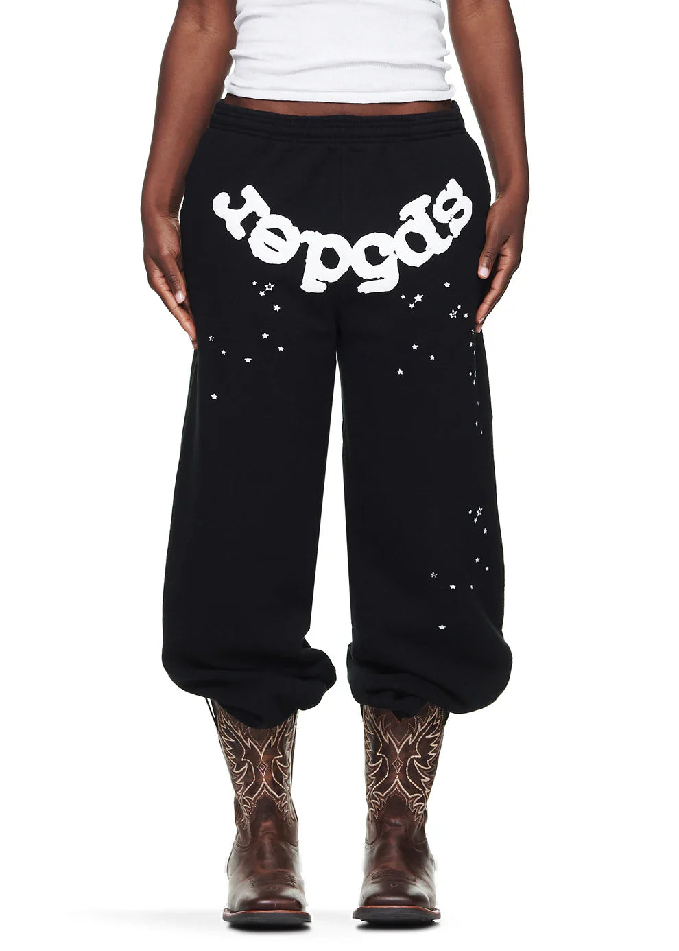 A person stands confidently against a white background, wearing Sp5der Sweatpants—unisex black cotton sweatpants with a bold front logo and star print—paired with a white tank top and brown embroidered cowboy boots.