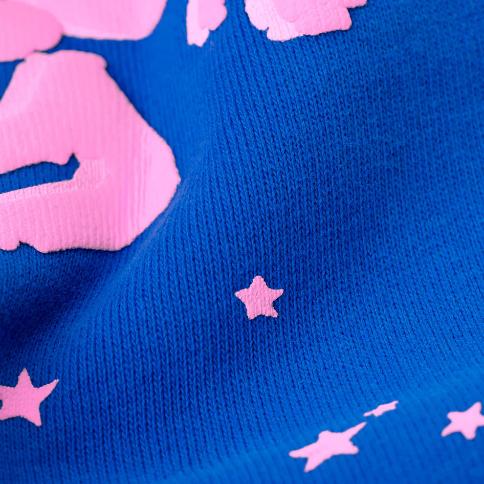 A close-up of the 100% Cotton Sp5der Sweatpants shows blue, ribbed material with pink logo and star accents, gentle folds, and a relaxed fit—perfect unisex jogger pants for trendy streetwear.
