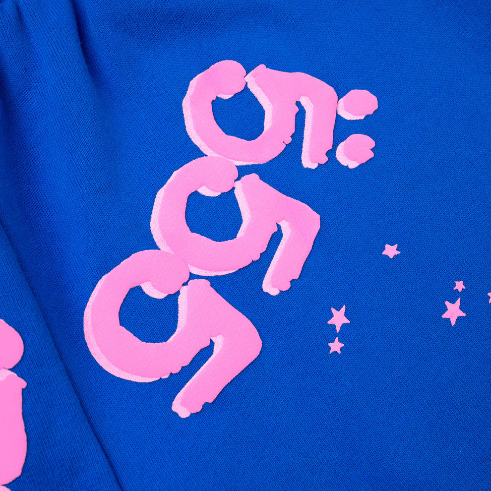A close-up of 100% Cotton Sp5der Sweatpants shows unisex blue jogger pants with pink logo and star accents, including stylized 9:59 numbers, highlighting their bold streetwear style and relaxed fit.